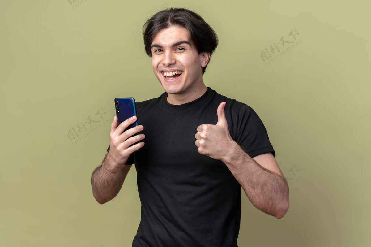 笑着笑容可掬的年轻帅哥 身穿黑色t恤 手持手机 大拇指向上竖立 孤零零地站在橄榄绿的墙上电话抱着衣服
