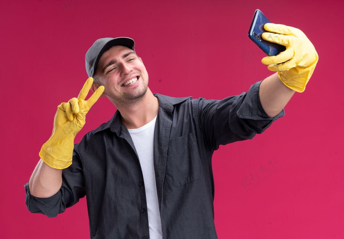 人带着微笑的年轻帅气的清洁工 穿着t恤 戴着手套 戴着帽子 在粉红色的墙上做一个显示和平姿态的自拍微笑脸感觉