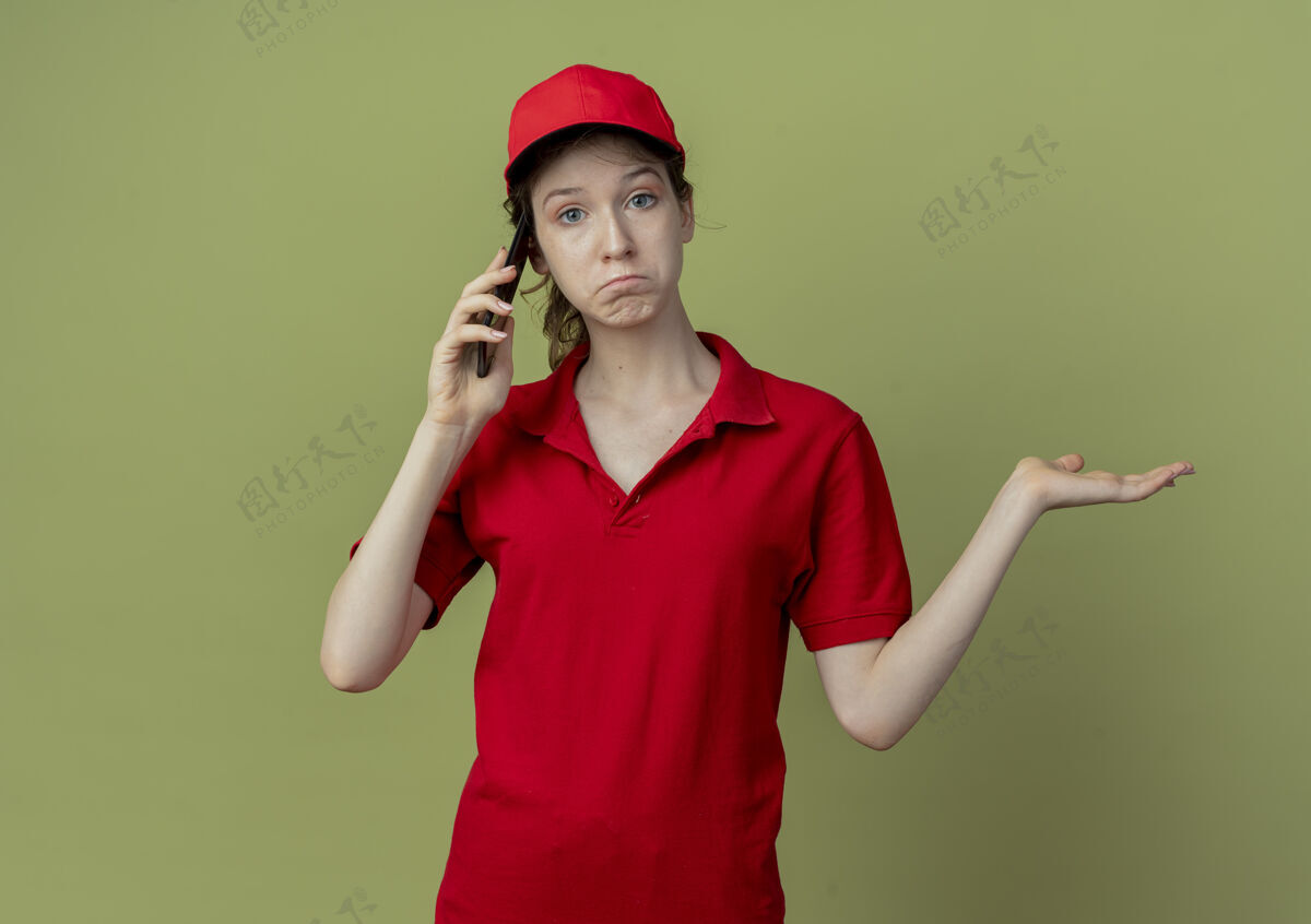 帽子困惑的年轻漂亮的送货女孩 穿着红色制服 戴着帽子 一边打电话 一边在橄榄绿的背景上展示着孤立的空手 还有复印空间交货漂亮年轻