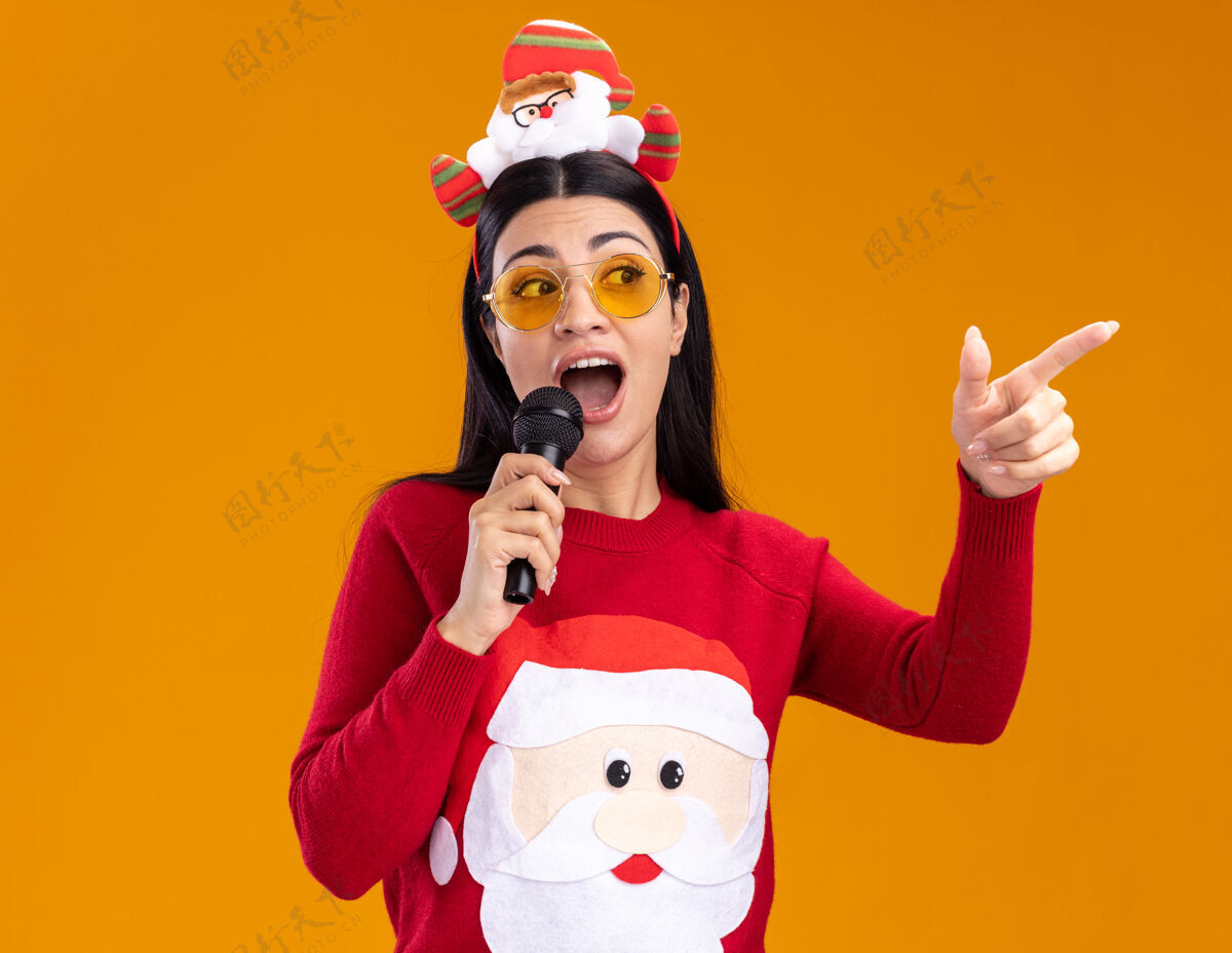 橙色给人印象深刻的年轻白人女孩戴着圣诞老人的头带和毛衣戴着眼镜对着麦克风说话看着并指着橙色背景上孤立的一面指向年轻圣诞老人