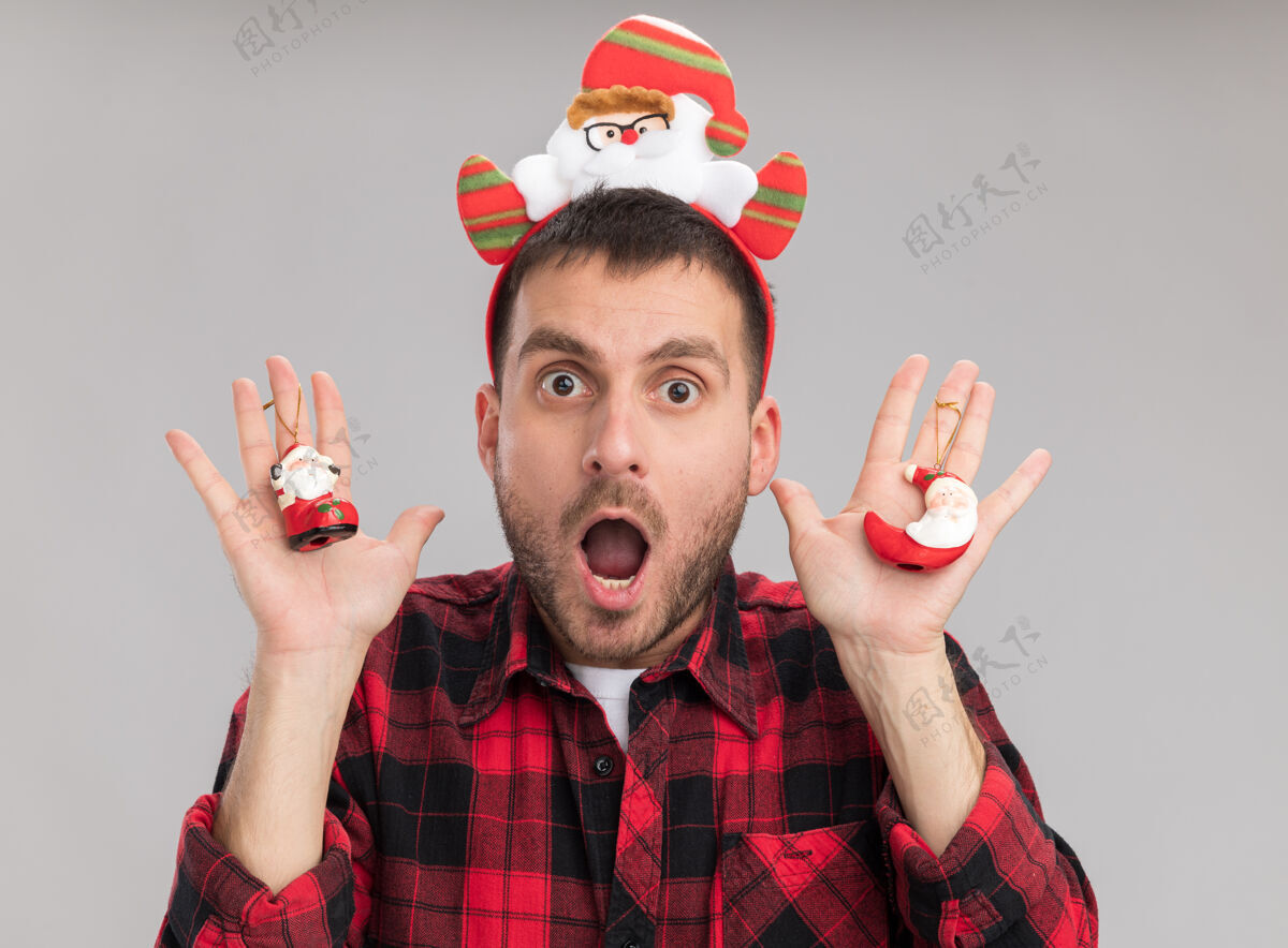 装饰品令人印象深刻的年轻白人男子戴着圣诞老人头带 手持圣诞老人的圣诞饰品 看着隔离在白色背景上的相机圣诞老人头带年轻