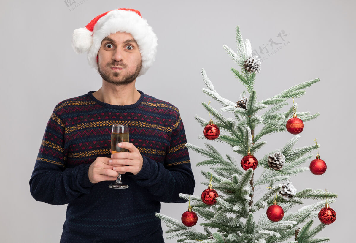 脸颊令人印象深刻的年轻白种人戴着圣诞帽站在圣诞树旁拿着一杯香槟看着相机喘气的脸颊孤立在白色背景帽子圣诞快乐男人
