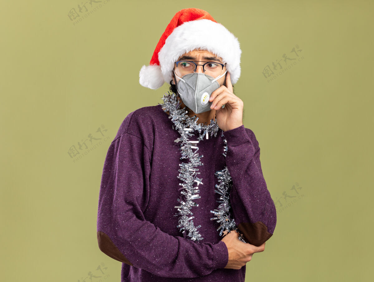 绿色一个年轻帅哥戴着圣诞帽 戴着医用面具 脖子上戴着花环 手放在脸颊上 在橄榄绿的背景下与世隔绝帽子圣诞快乐圣诞