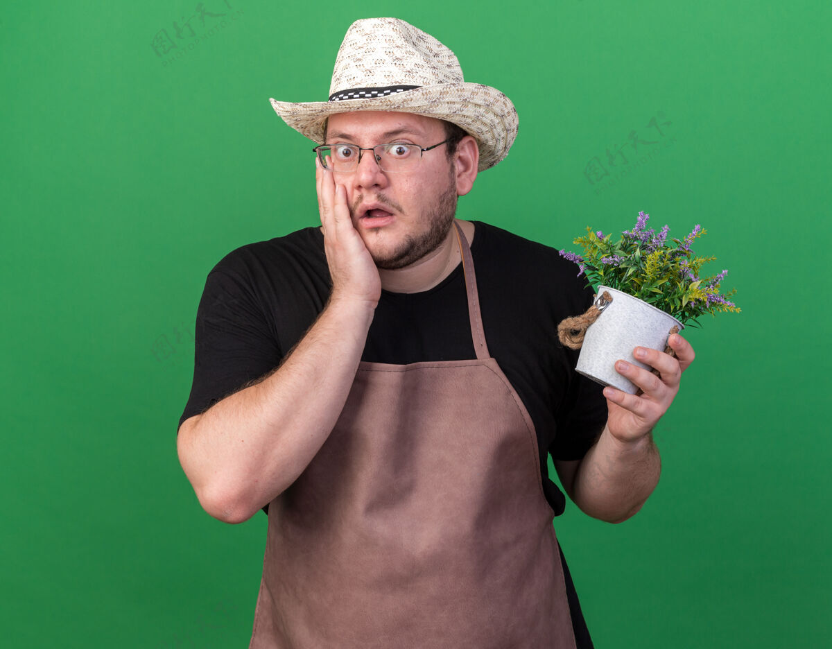 园艺年轻的男园丁戴着园艺帽 手里拿着花盆里的花 手放在隔离在绿墙上的脸颊上 吓得要命人脸颊姿势