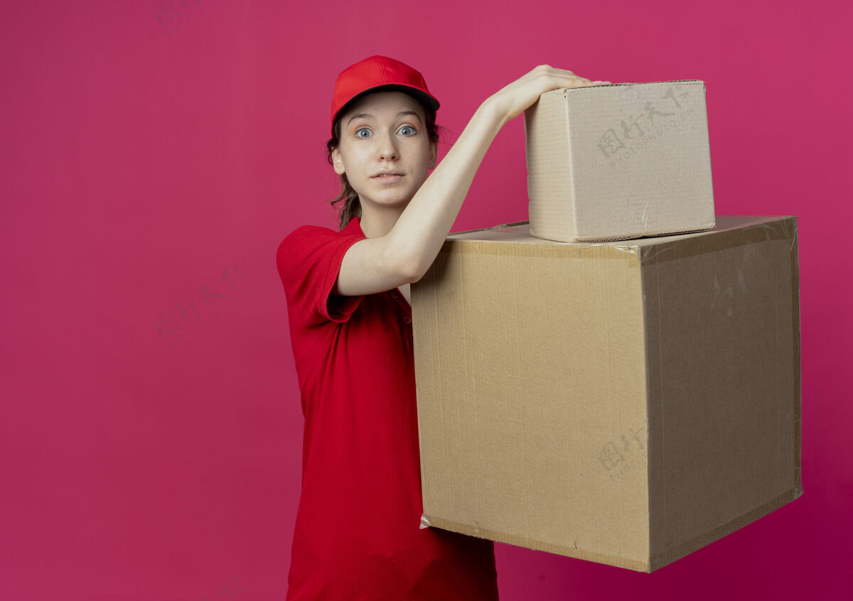 红色令人印象深刻的年轻漂亮的送货女孩在红色制服和帽子举行纸箱看着摄像机在深红色的背景与复制空间隔离背景持有帽子