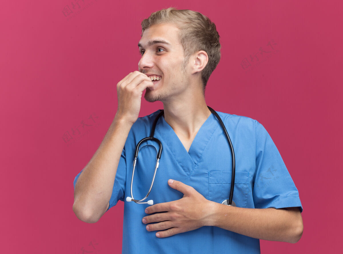 穿微笑着看着身边年轻的男医生穿着医生制服用听诊器咬指甲隔离在粉红色的墙上市民壁板男性