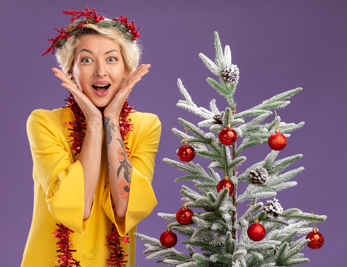 花圈兴奋的金发女郎头戴圣诞花环 脖子上戴着金属丝花环 站在装饰好的圣诞树旁 手放在脸上 看着紫色背景上孤立的摄像机头圣诞脖子