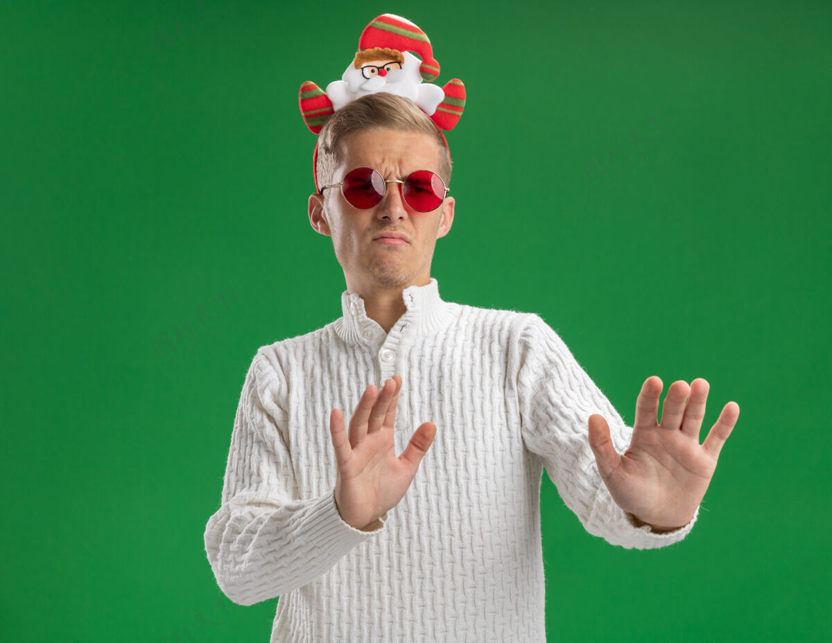 空间愁眉苦脸的年轻帅哥戴着圣诞老人的头带 戴着眼镜 看着一边做着拒绝的手势 隔离在绿色的背景上头带皱眉绿色