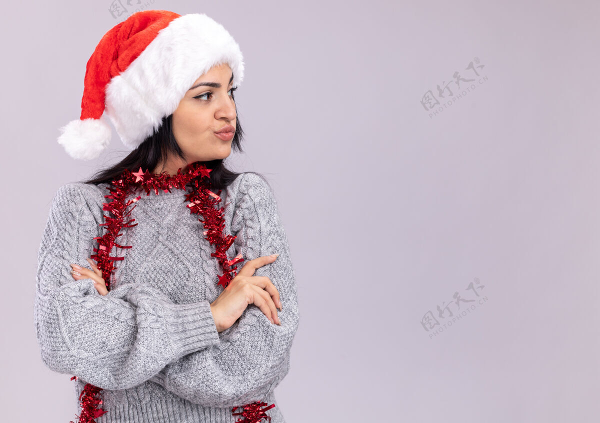 立场可疑的年轻白人女孩戴着圣诞帽 脖子上戴着金箔花环 站在白色背景下 以封闭的姿势看着孤立的一面脖子白色圣诞快乐