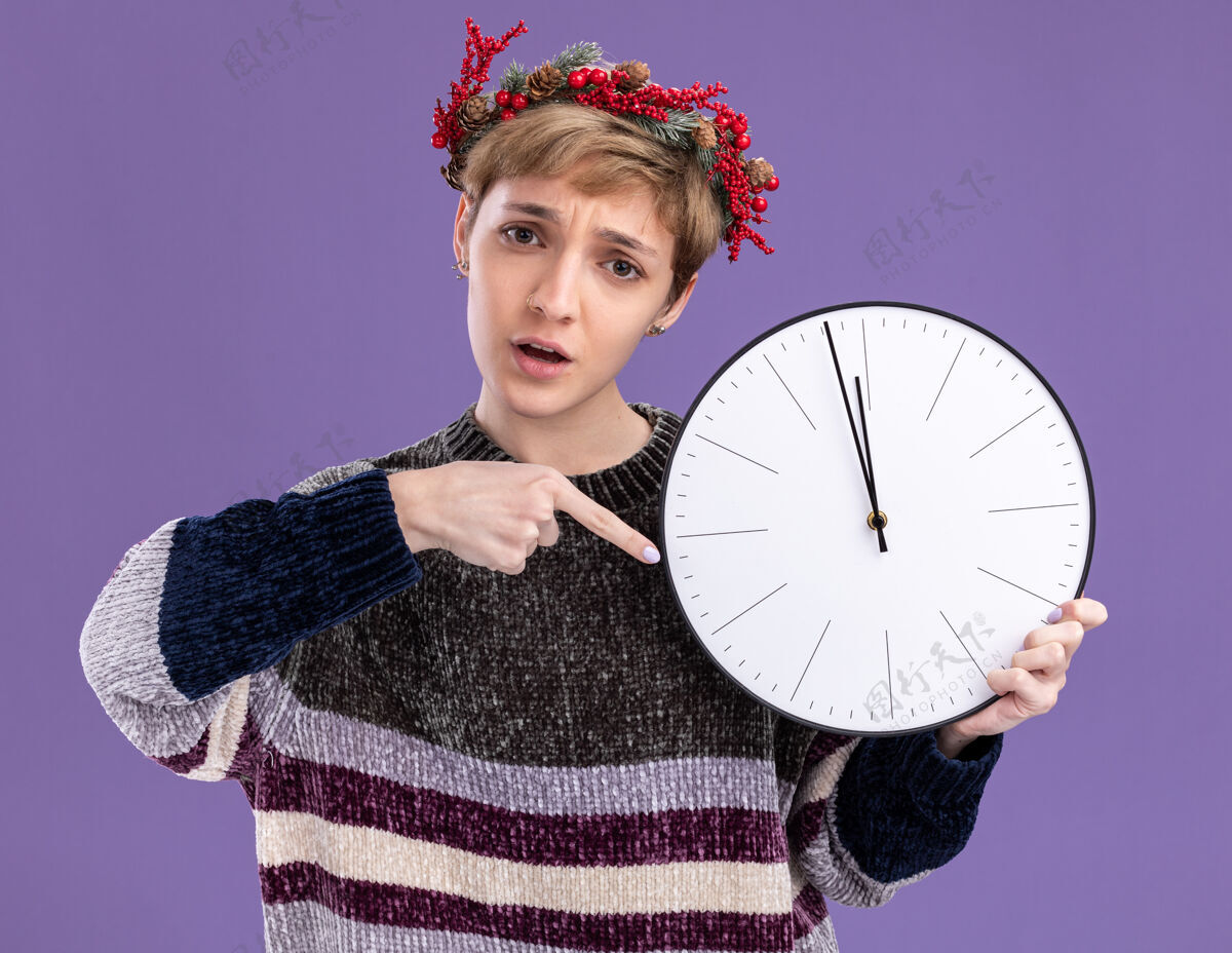 圣诞快乐焦急的年轻漂亮女孩戴着圣诞花环 手持并指着时钟 看着隔离在紫色背景上的相机抱着花圈紫色