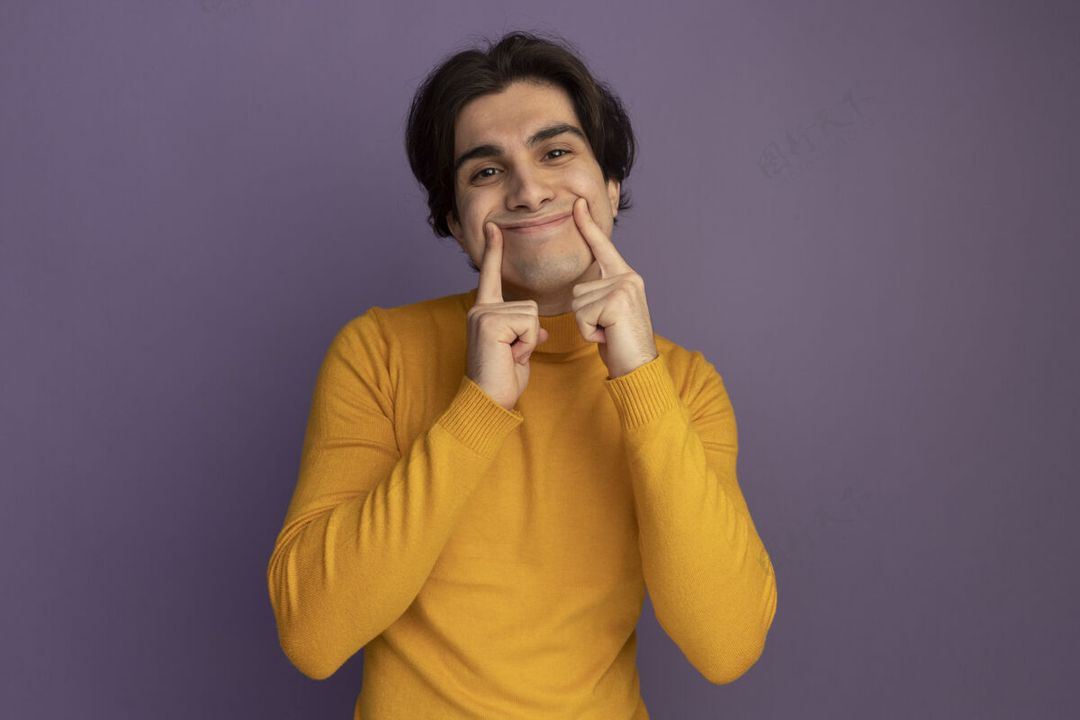 帅哥高兴的年轻帅哥穿着黄色高领毛衣做微笑 手指孤立在紫色的墙上手指高领毛衣男人