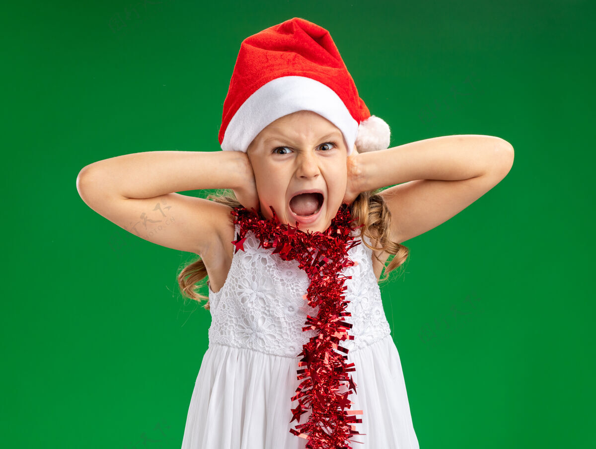 小恼怒的小女孩戴着圣诞帽 脖子上戴着花环 耳朵被隔离在绿色背景上圣诞快乐圣诞节女孩