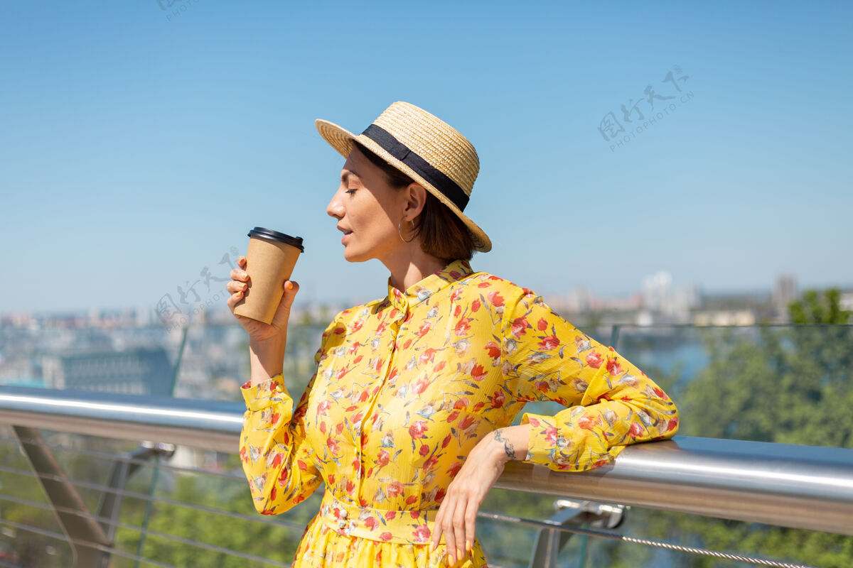 咖啡穿着黄色夏装 戴着黄色帽子 喝着咖啡 享受阳光的女人的户外肖像 站在桥上 城市的景色令人惊叹20多岁旅游自信