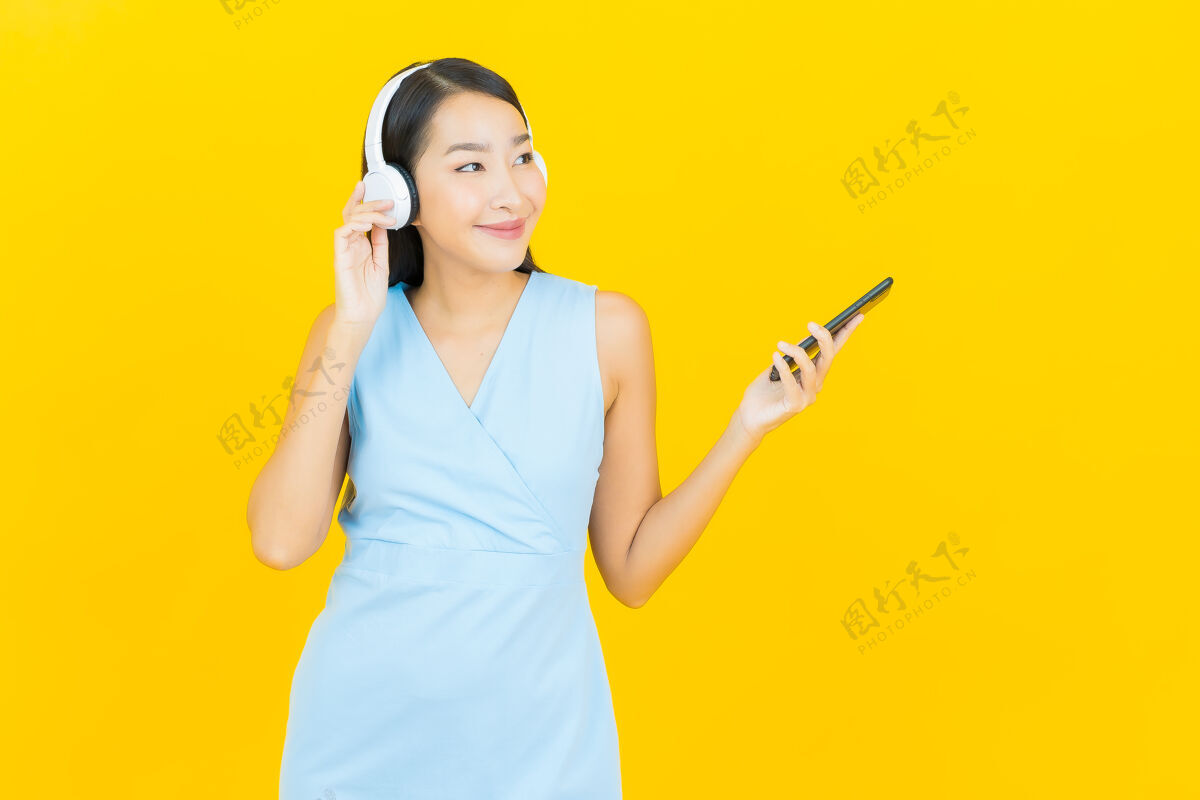 成人画像美丽的亚洲年轻女子戴着耳机和智能手机在黄色的墙上听音乐女性年轻脸