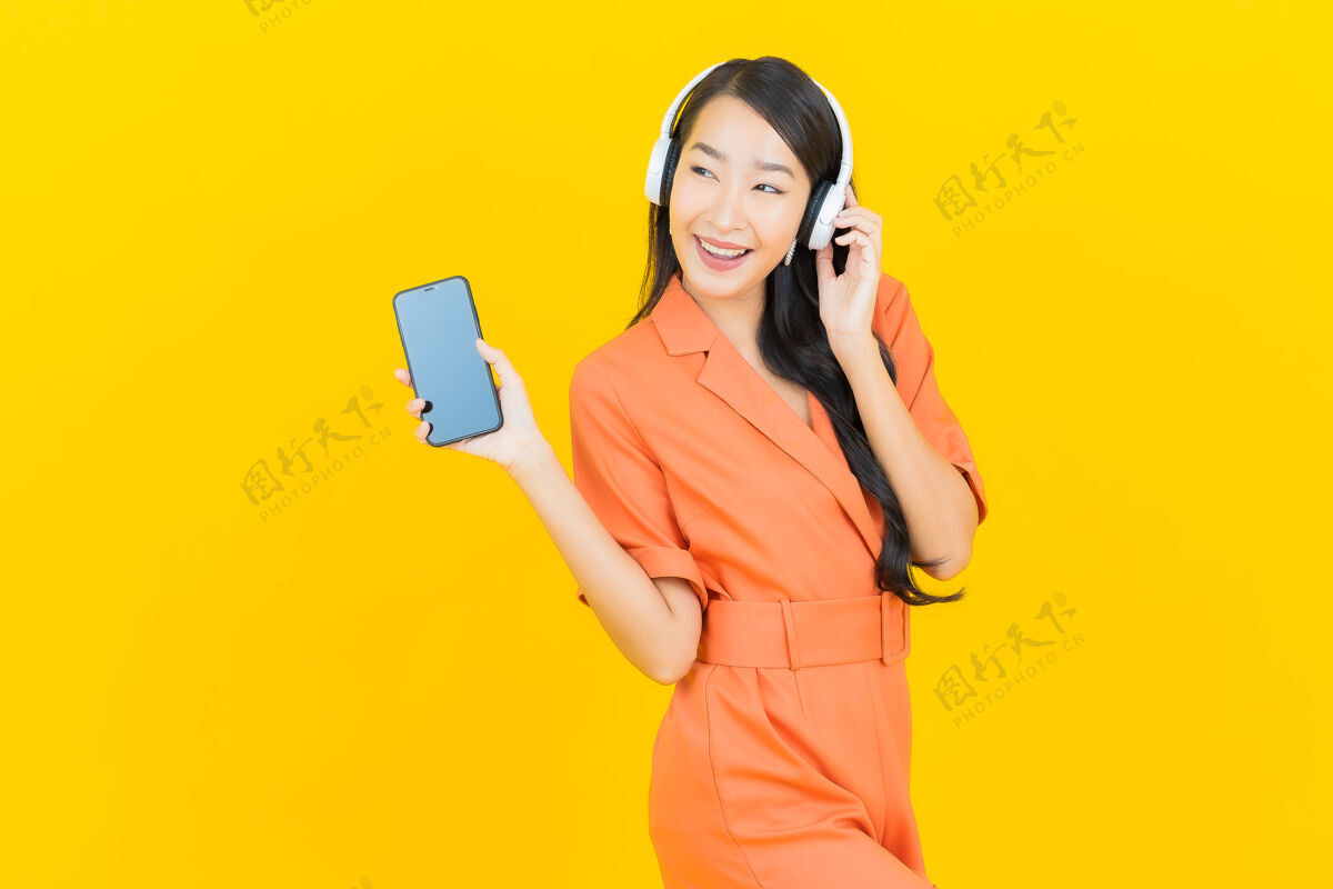 收音机肖像美丽的亚洲年轻女子戴着耳机和智能手机听黄色音乐橙色连身裤黄色肖像