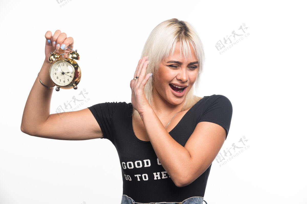 成人在白色的墙上 年轻的女人拿着钟 手指向钟女人金发表达