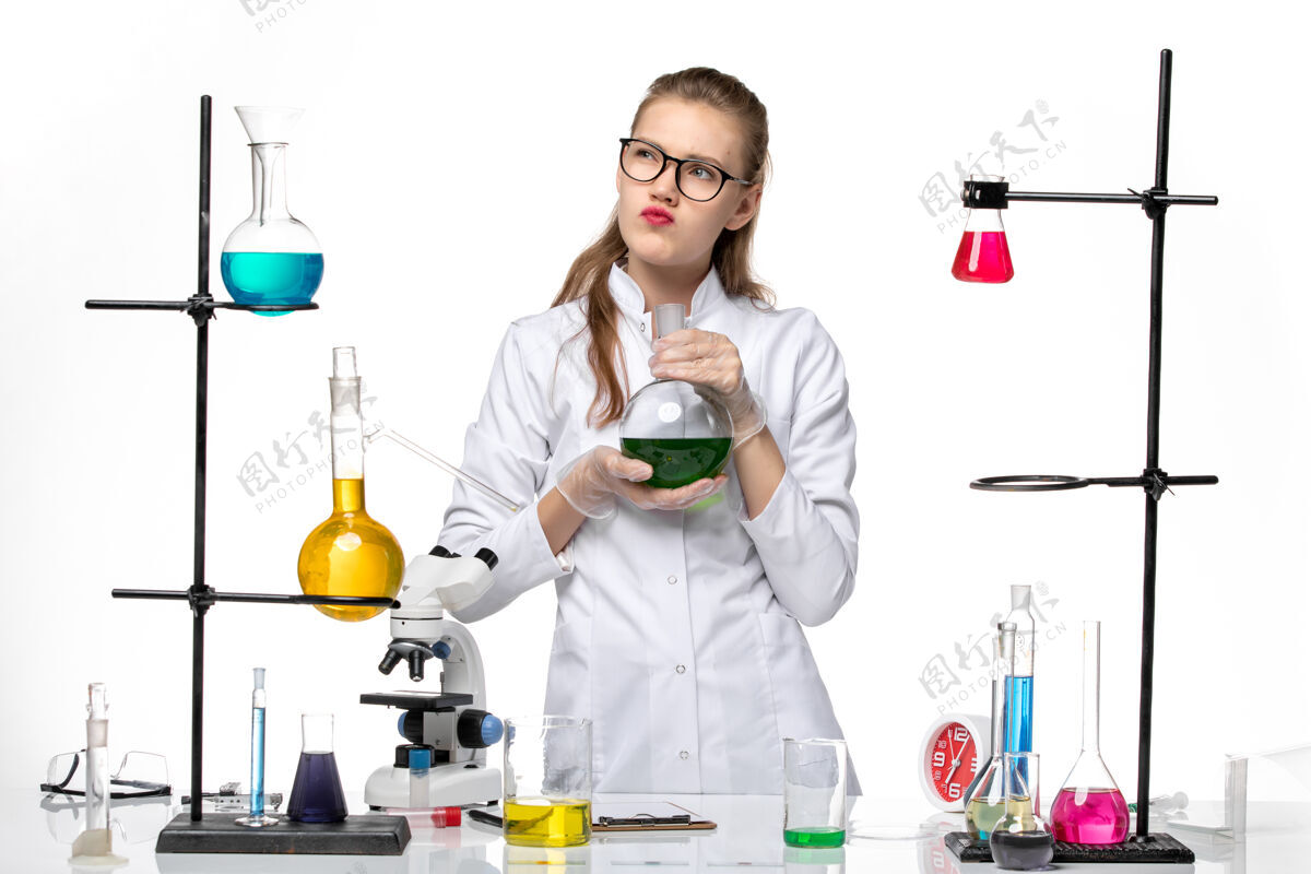化学前视图穿着医疗服的女化学家拿着装有绿色溶液的烧瓶在白色背景上化学大流行的病毒观点绿色前面