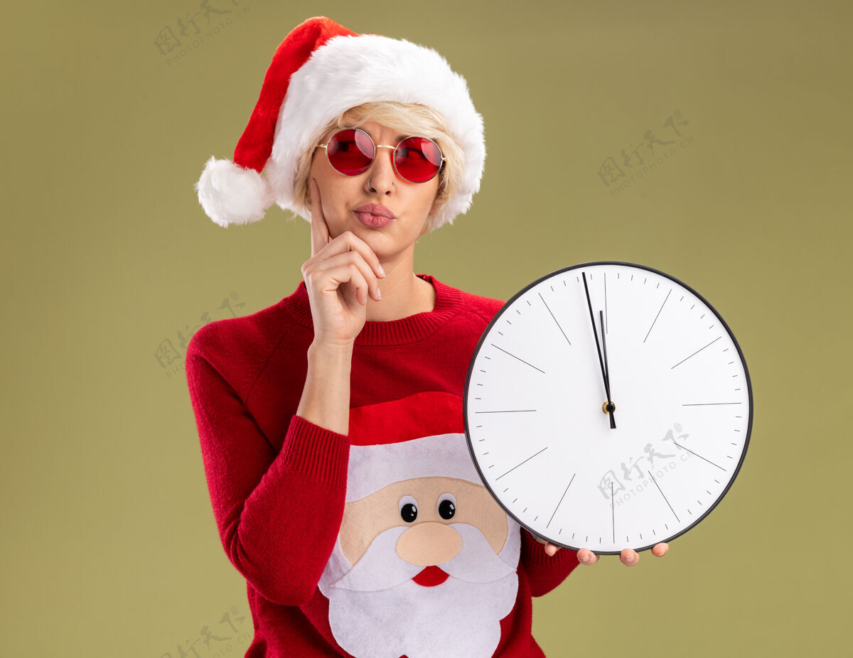 眼镜可疑的年轻金发女人戴着圣诞帽和圣诞老人的圣诞毛衣戴着眼镜拿着时钟手放在下巴上看着橄榄绿背景上孤立的一面怀疑圣诞金发