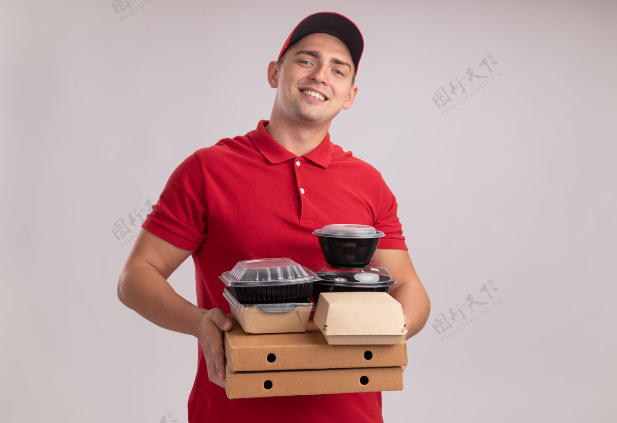 食物面带微笑的年轻送货员穿着制服 戴着帽子 把食物容器放在隔离在白墙上的比萨饼盒上人制服姿势