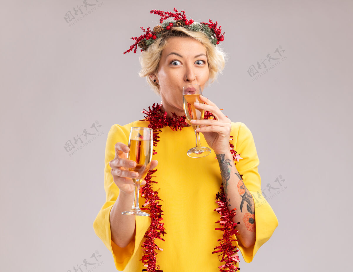 头年轻的金发女郎戴着圣诞花环 脖子上戴着金箔花环 手里拿着两杯香槟 看着相机 一边喝着一杯 背景是白色的二眼镜空间