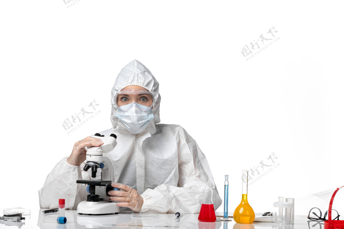 显微镜前视图：女医生穿着防护服 戴着口罩 在白色背景下使用显微镜观察病毒大流行时溅起的病毒药喷溅白色