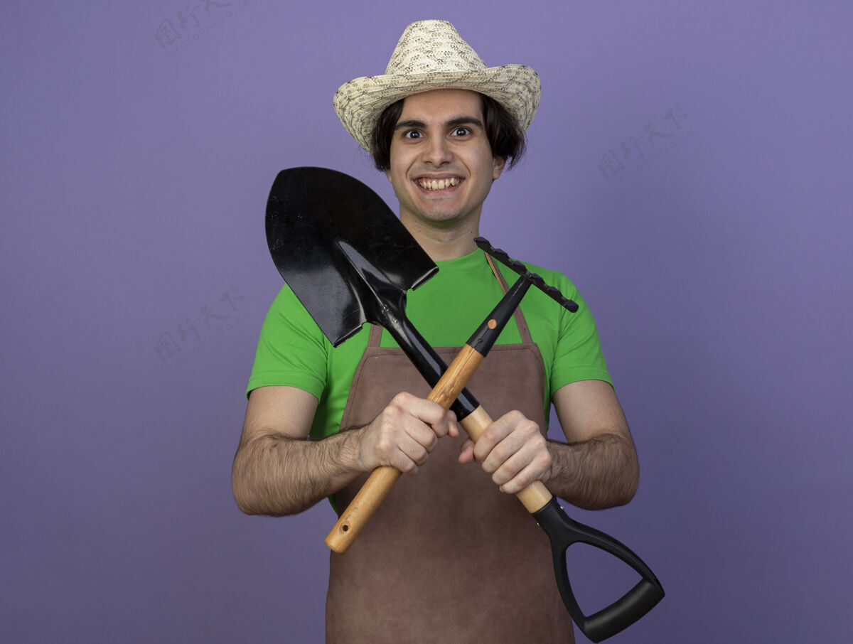 人微笑着的年轻男园丁 穿着制服 戴着园艺帽 拿着铲子穿过耙子男人花园年轻