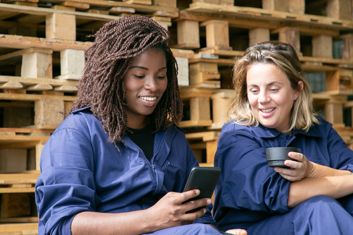 观看快乐的工厂同事们穿着工作服一起看手机上的内容 一边在仓库里喝咖啡中年聊天手机