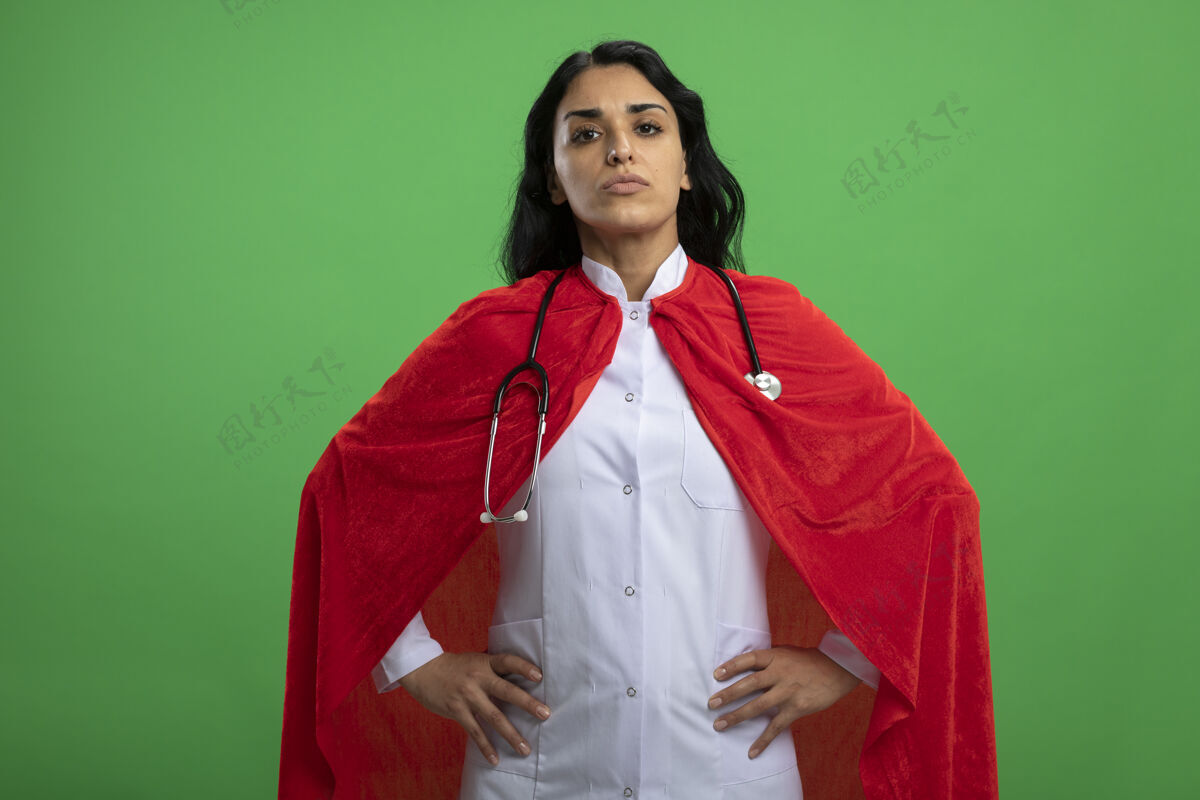 穿自信的年轻超级英雄女孩穿着医用长袍 手放在臀部 隔离在绿墙上手听诊器医疗
