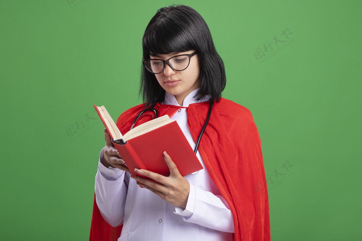 年轻年轻的超级英雄女孩戴着听诊器 穿着医用长袍 披着斗篷 戴着眼镜 拿着书看着隔离在绿墙上的书超级英雄医疗穿