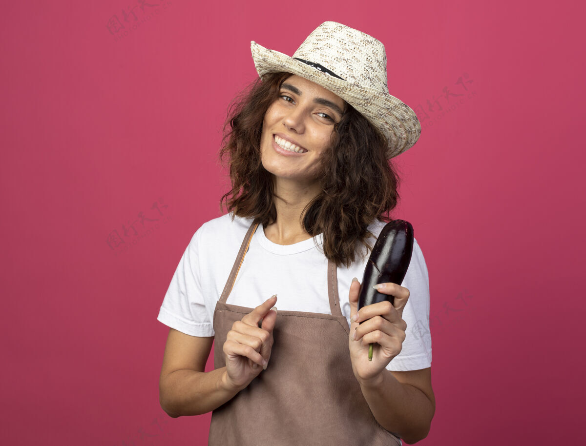 点年轻的女园丁面带微笑 穿着制服 戴着园艺帽 指着隔离在粉红色上的茄子园艺花园穿