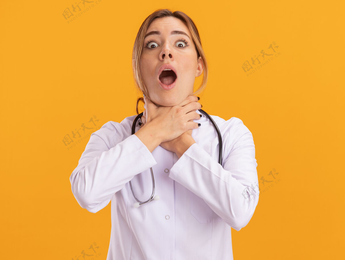 年轻年轻的女医生穿着带听诊器的医用长袍 抓住隔离在黄墙上的喉咙穿长袍听诊器