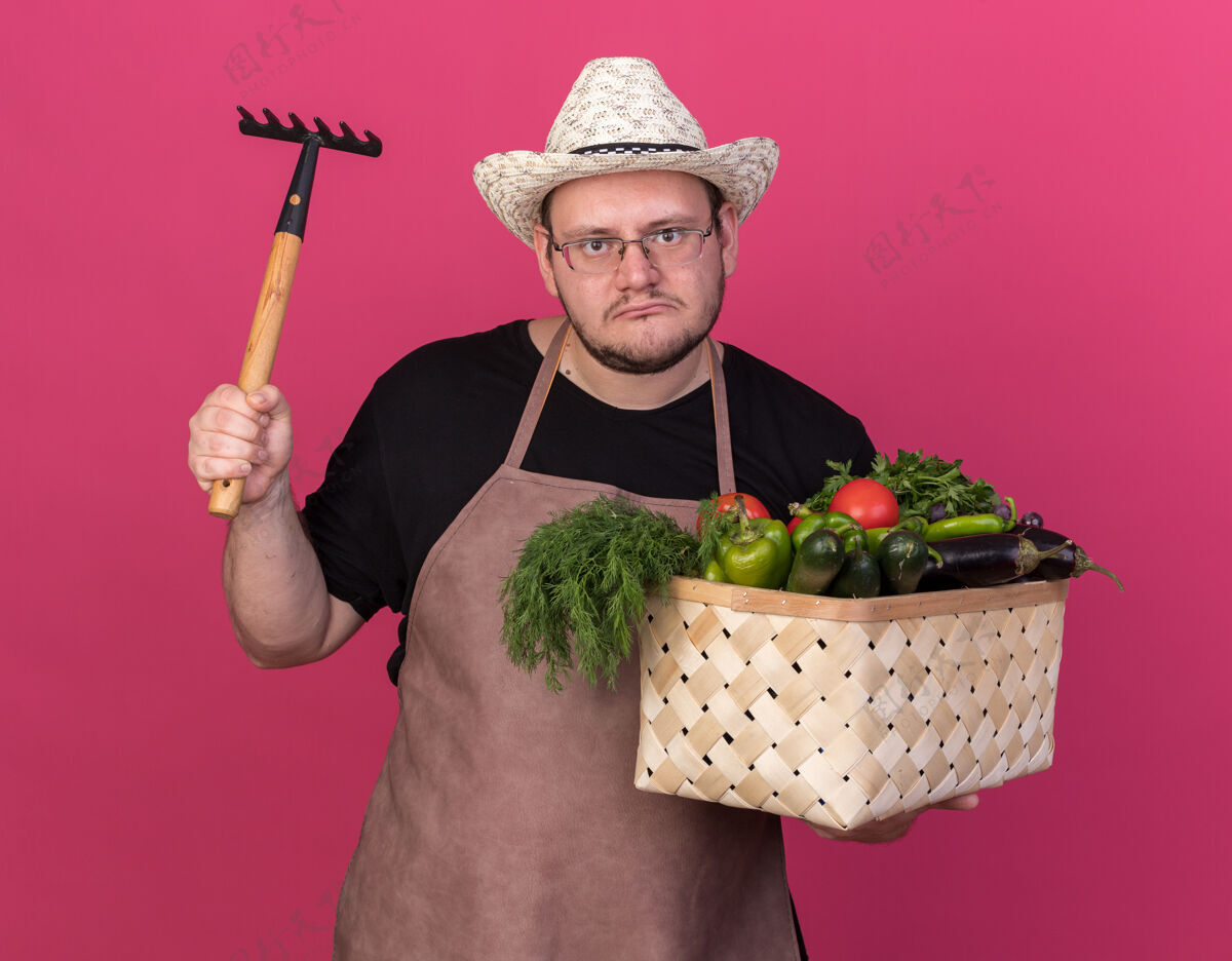 蔬菜年轻的男园丁戴着园艺帽 拿着菜篮 把耙子孤立在粉红色的墙上提高持有花园