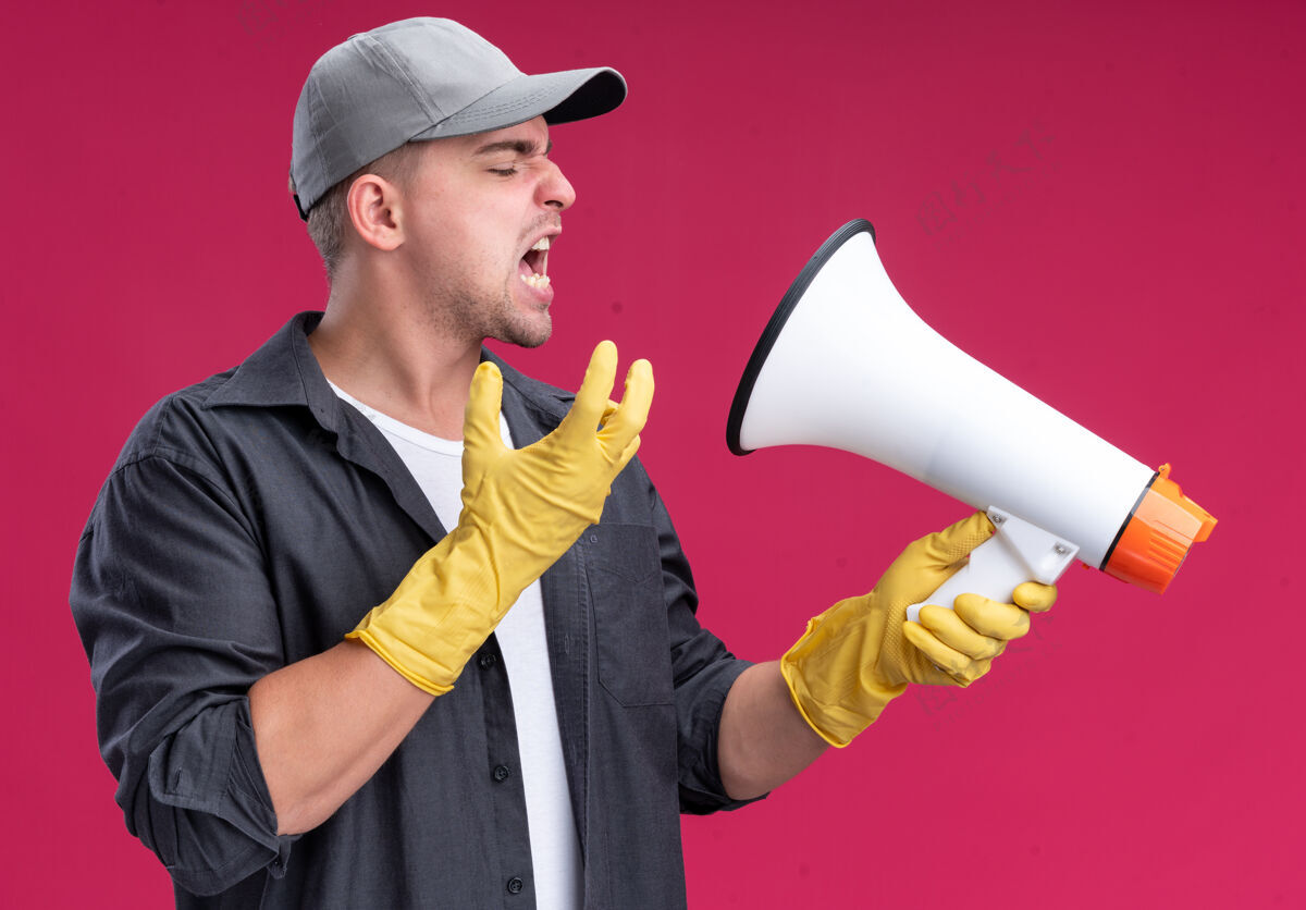 愤怒愤怒的年轻汉姆森保洁员穿着t恤 戴着手套戴着帽子 在隔离在粉红色墙上的扬声器上讲话年轻清洁穿
