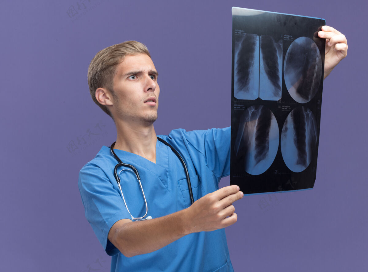 戴害怕的年轻男医生穿着医生制服手持听诊器 看着隔离在蓝色墙上的x光片听诊器男性医生