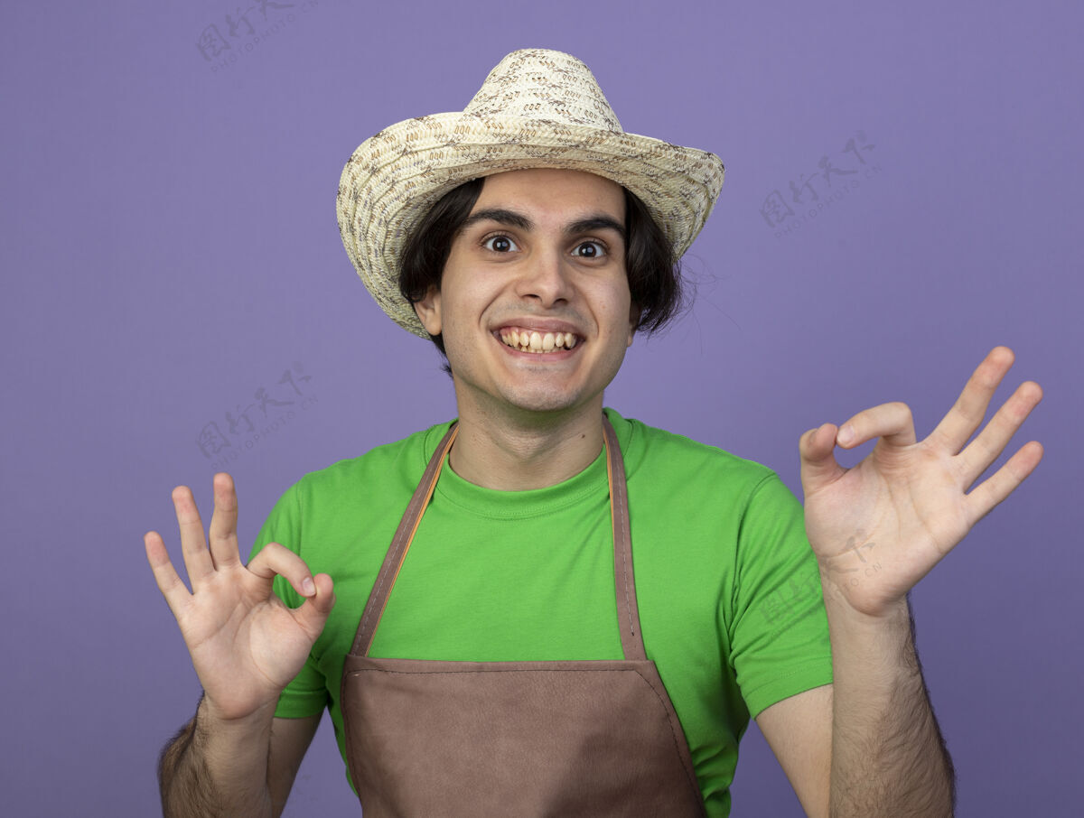 伙计微笑着的年轻男园丁穿着制服 戴着园艺帽 在紫色上显示出良好的姿态人们好的手势