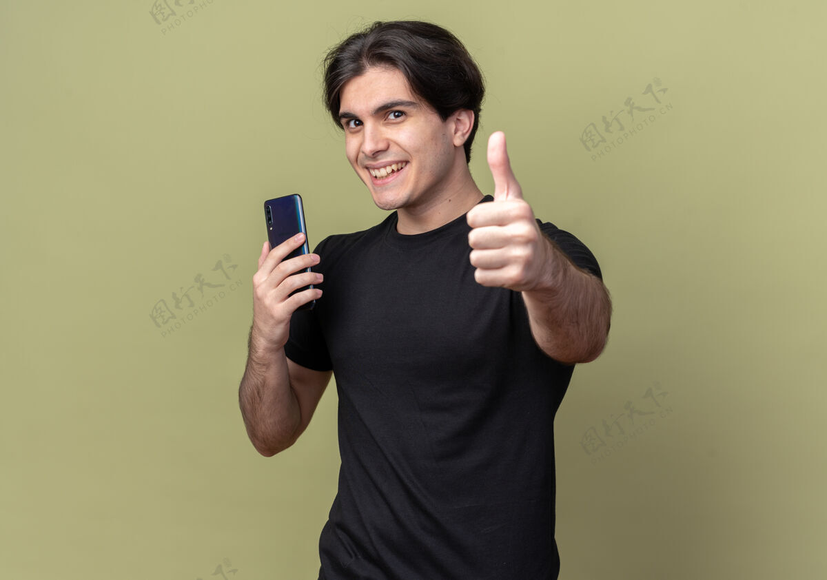 秀带着微笑的年轻帅哥穿着黑色t恤拿着手机竖起大拇指孤立地站在橄榄绿的墙上 留着复印空间拇指微笑奥利弗
