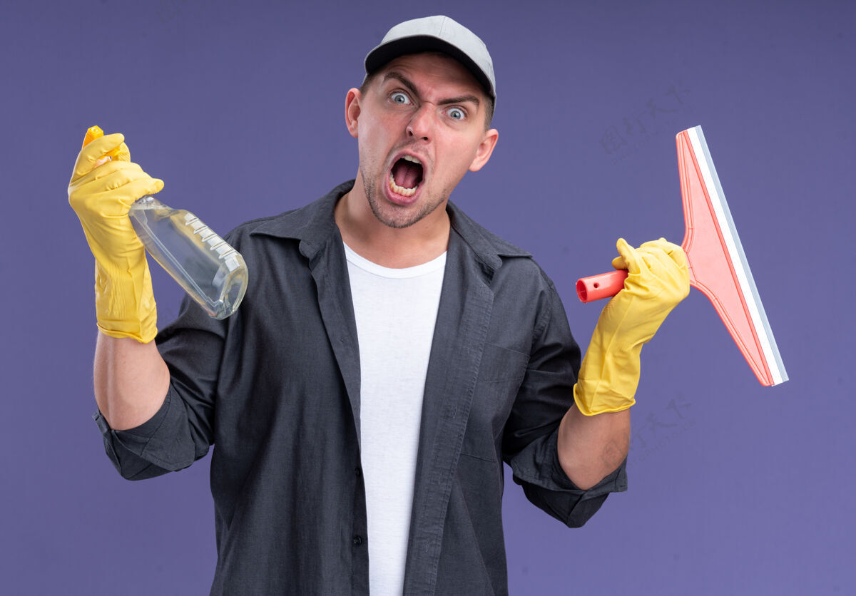 手套愤怒的年轻帅气的清洁工穿着t恤 戴着帽子和手套 拿着拖把头和喷雾瓶隔离在紫色的墙上喷雾穿举行