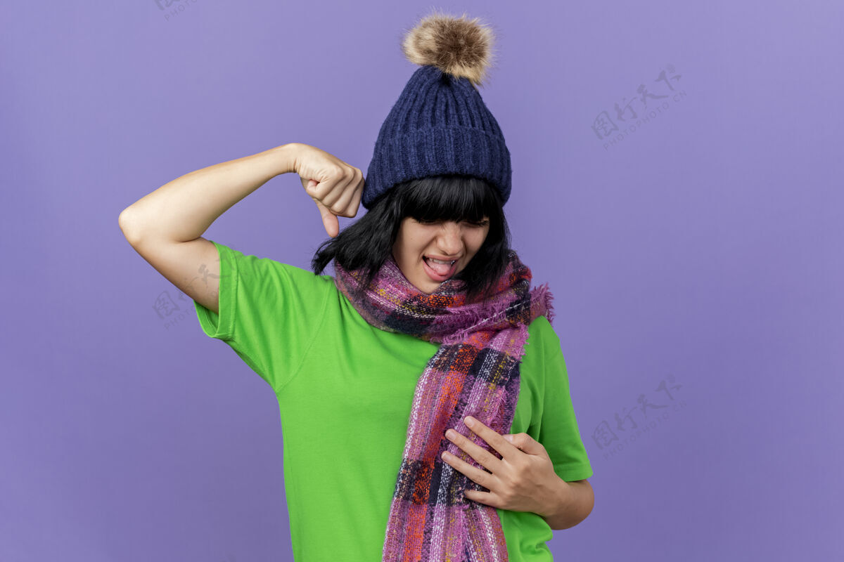人带着冬天帽子和围巾的疼痛的年轻生病的女人 手放在肚子上 拳头举在空中 闭着眼睛 隔离在紫色的墙上 留着复制空间脸拳头人