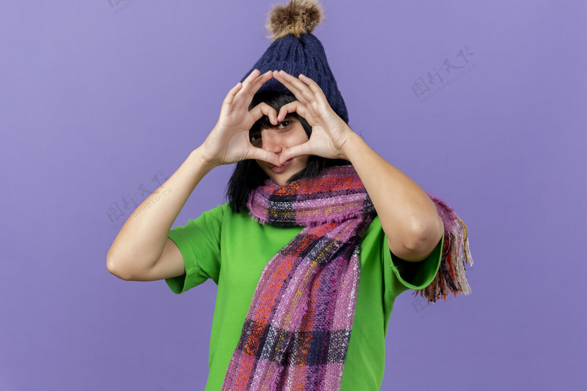 帽子年轻的生病的女人戴着冬天的帽子和围巾做着心形的招牌 透过它看前方 隔离在紫色的墙上 留着复制的空间人表情感情