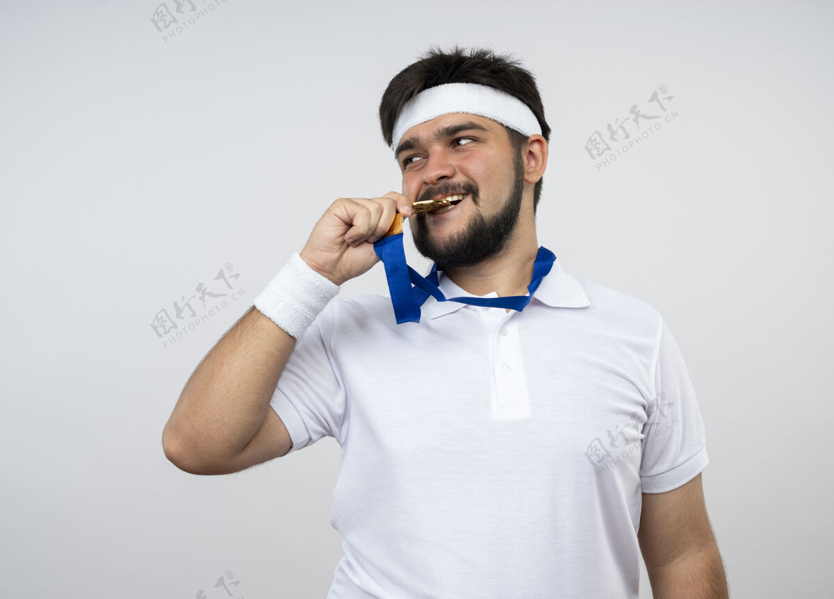 头带高兴的年轻人看着戴着头带 戴着手环的侧面 咬着隔离在白墙上的奖牌运动年轻奖牌
