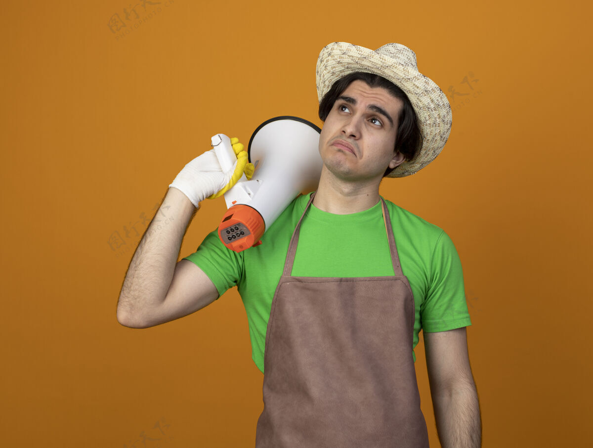 橙色悲伤的年轻男园丁穿着制服戴着园艺帽戴着手套看着一边拿着喇叭在肩膀上孤立的橙色穿壁板悲伤