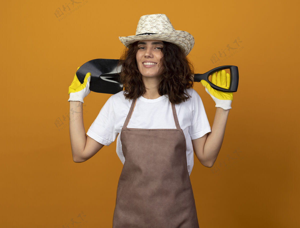 女人微笑着的年轻女园丁穿着制服戴着园艺帽和手套把铁锹放在脖子后面微笑帽子脖子