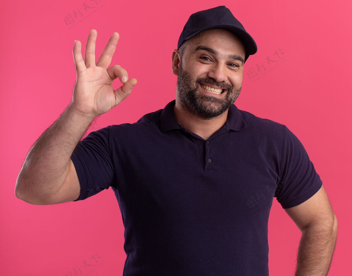 中年微笑的中年送货员穿着制服 戴着帽子 在粉红色的墙上显示出良好的姿态微笑男人好的