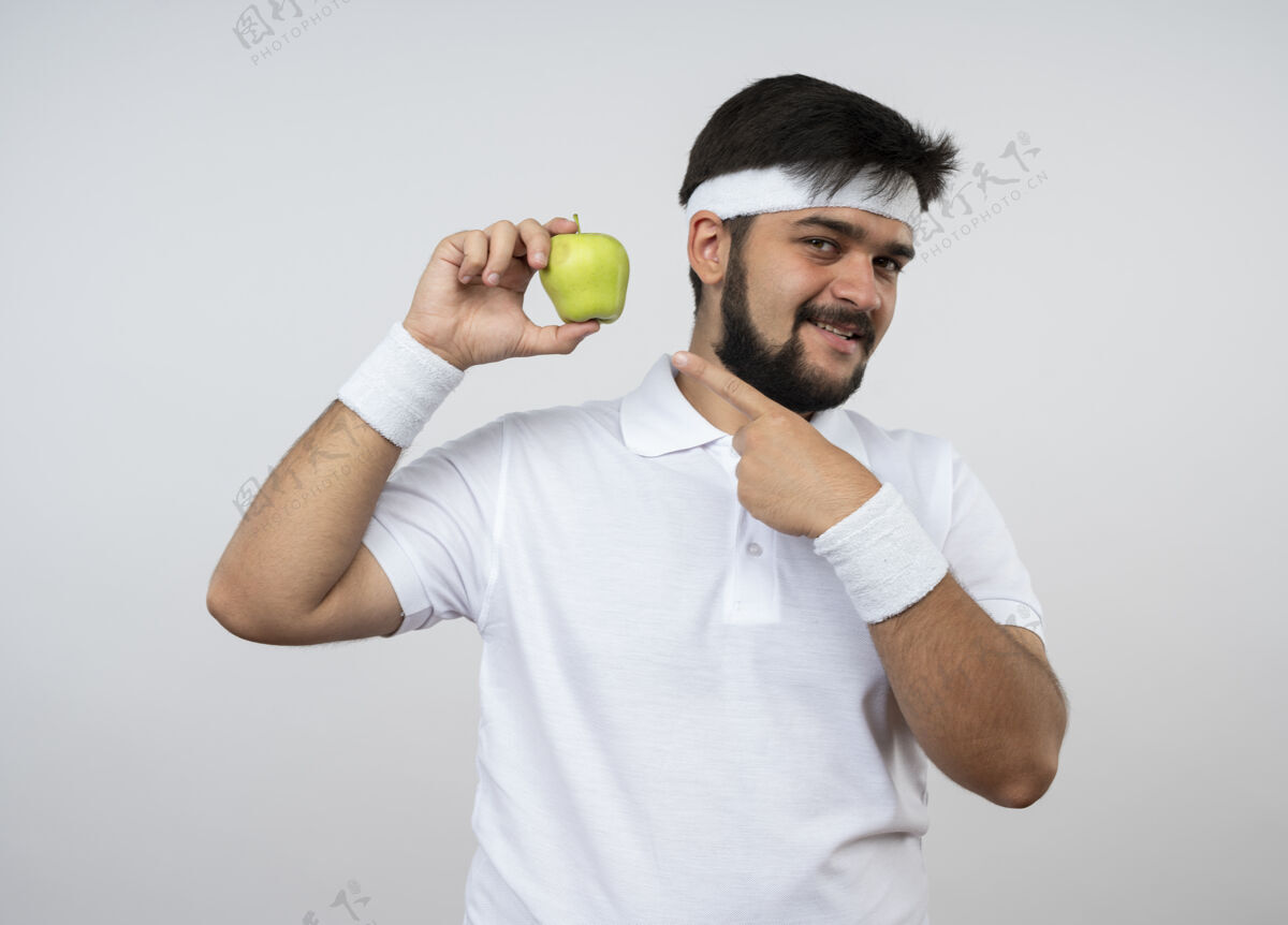 头带微笑着的年轻人戴着头带和腕带 指着隔离在白墙上的苹果点苹果年轻