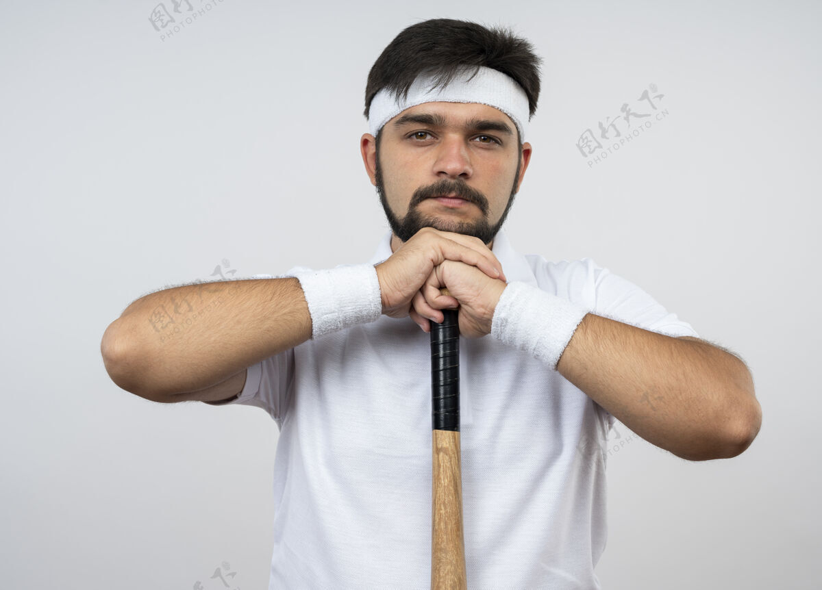 男人自信的年轻人戴着头带和腕带把下巴放在棒球棒上隔离在白墙上年轻腕带穿