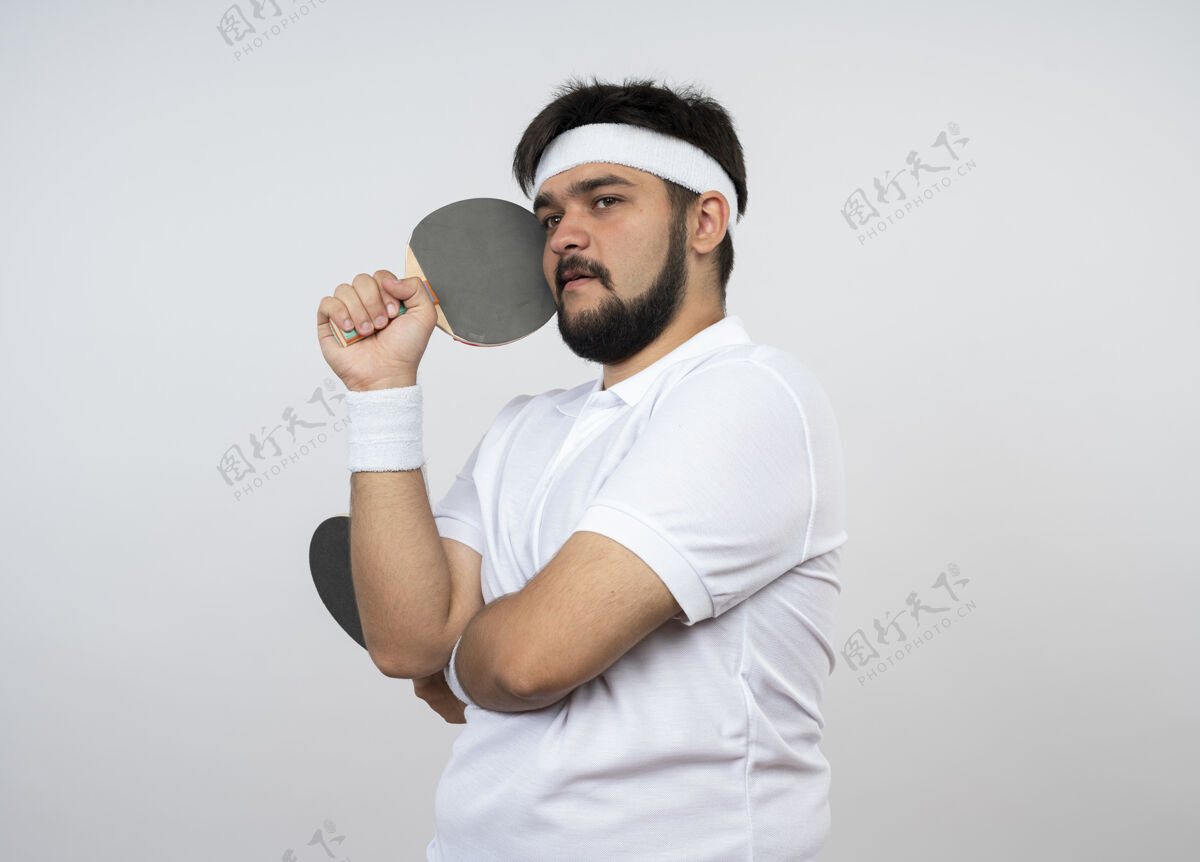 运动想着年轻的运动型男人 一边戴着头带 一边拿着乒乓球拍看着隔离在白墙上的脸乒乓球腕带抱
