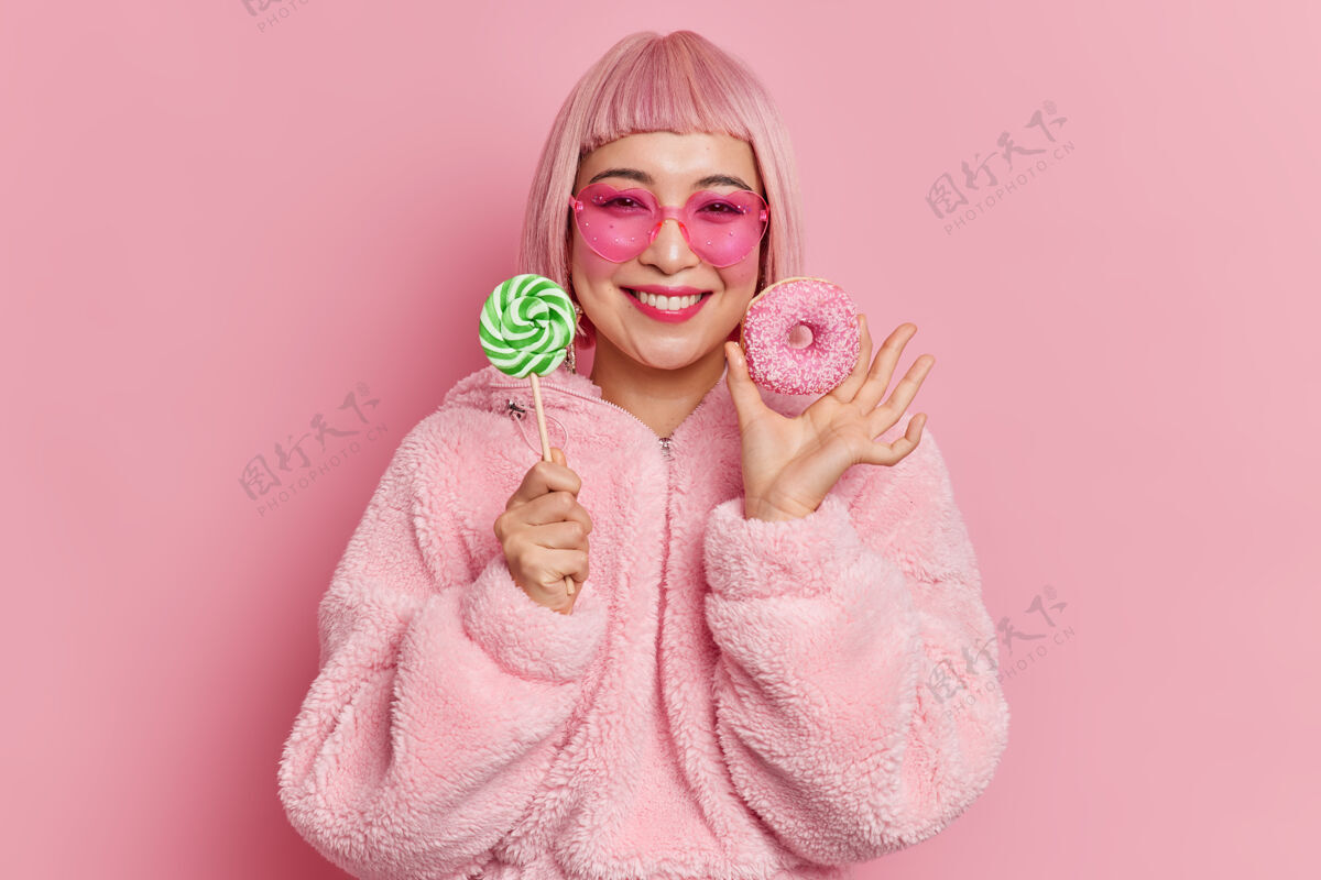 年轻满头粉红卷发的亚洲年轻女子微笑着 手里拿着棒棒糖和美味的甜甜圈外套人亚洲
