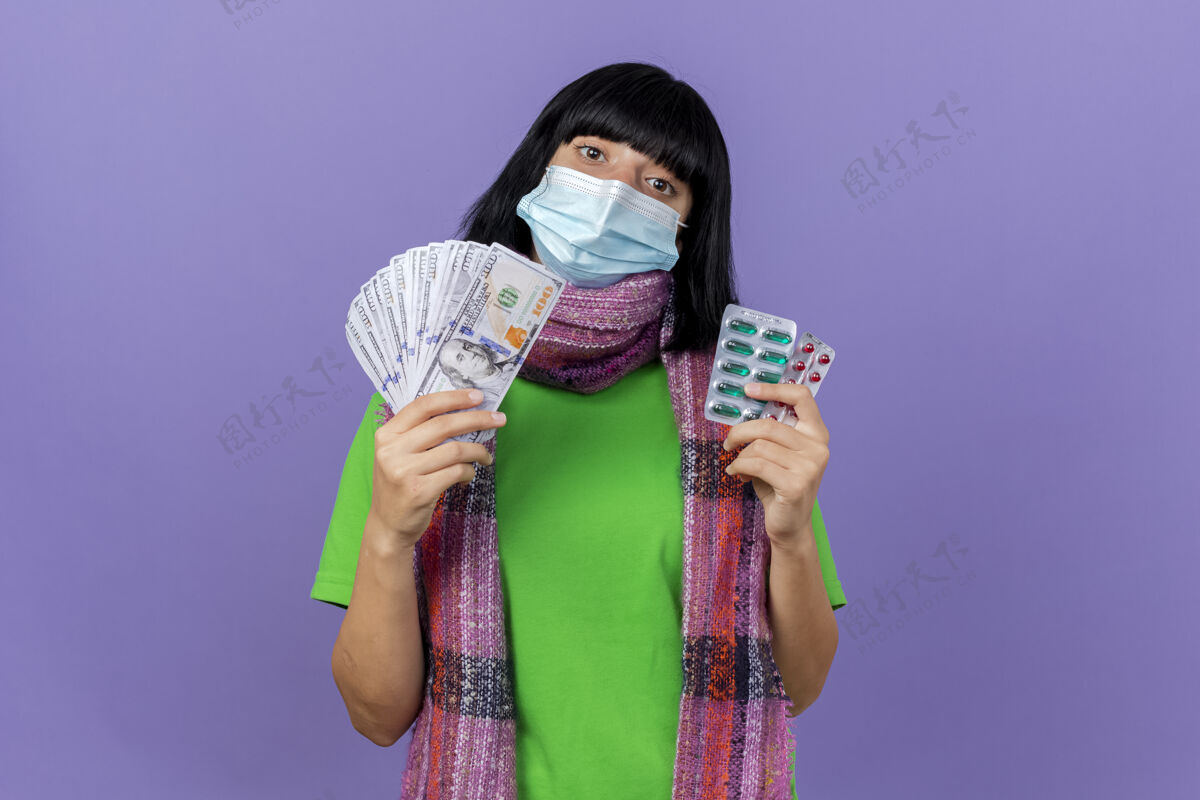 人年轻生病的女人戴着口罩 戴着围巾 手里拿着钱和一包包胶囊 看着前面隔离着的紫色墙壁上有复印空间包疾病人