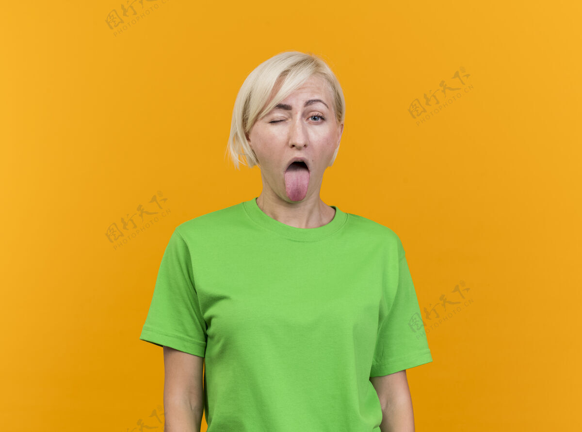 站着顽皮的中年金发斯拉夫女人在看前面显示舌头隔离在黄色墙上与复制空间玩舌头金发