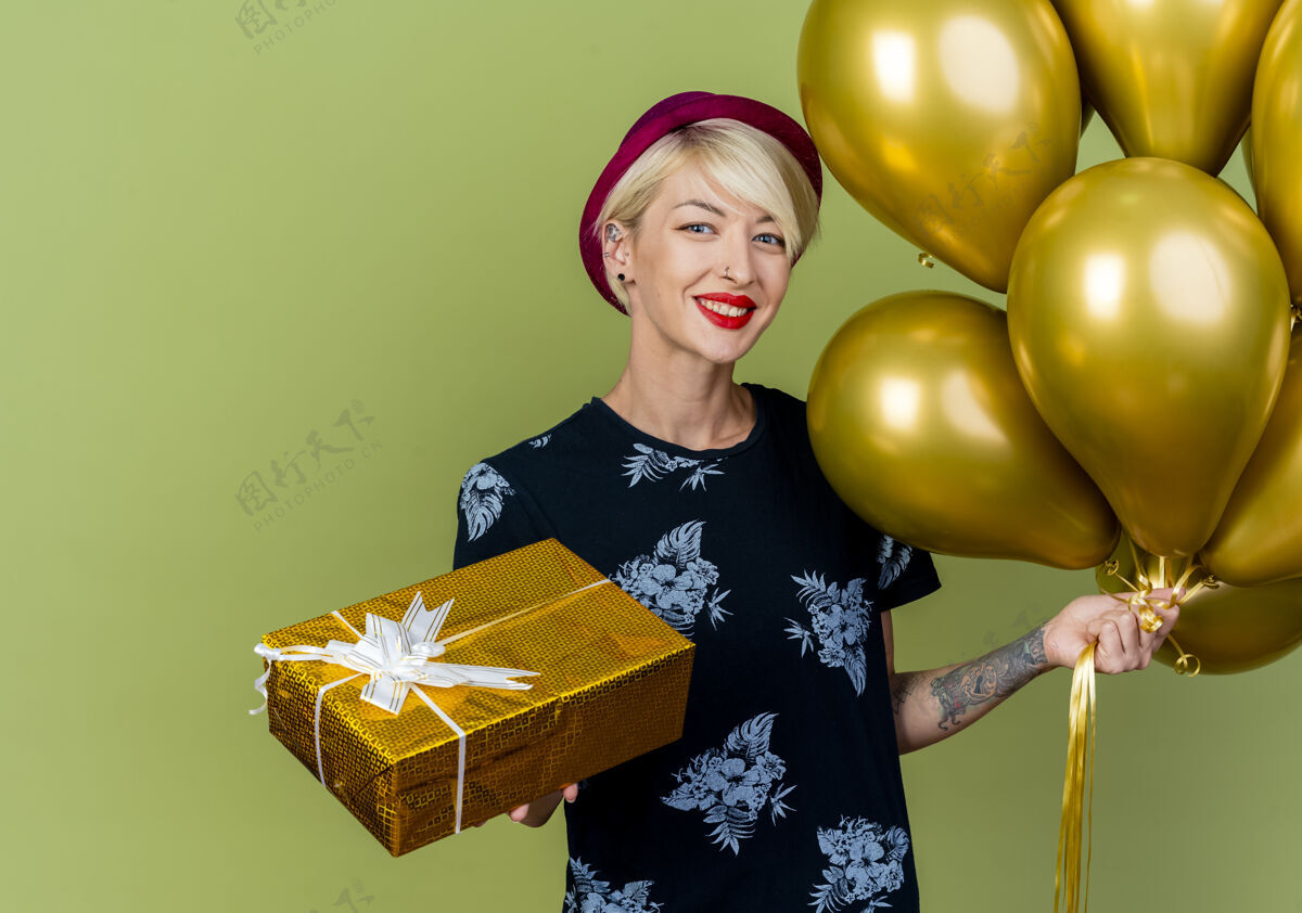 黄金微笑着的年轻金发派对女郎戴着派对帽拿着气球和礼盒看着橄榄绿墙上孤立的前方表情站着黄金